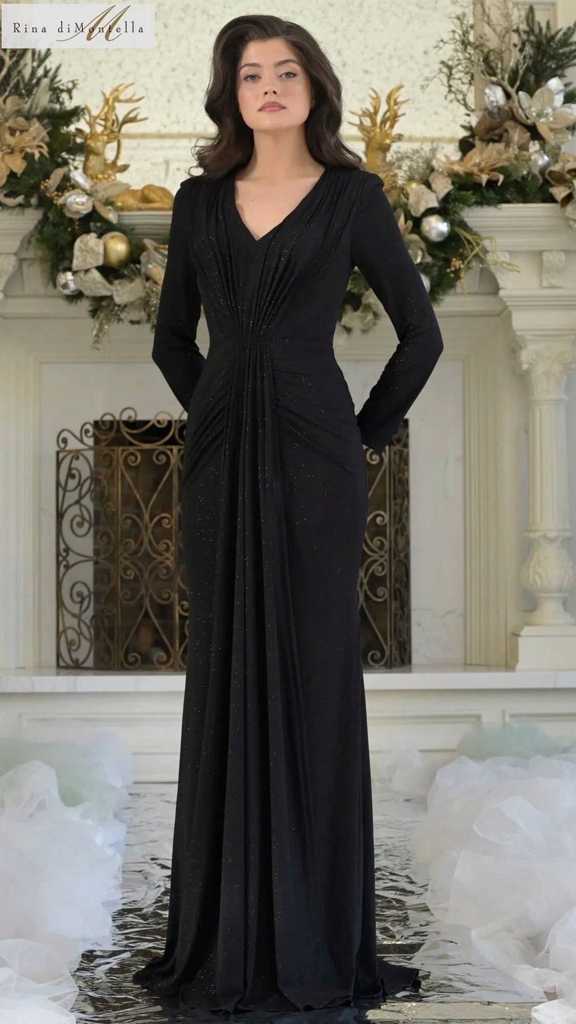 A woman stands indoors in front of a decorated fireplace wearing the black Rina Di Montella RD3348 long formal dress, featuring gathered detailing and surrounded by white and gold floral arrangements.